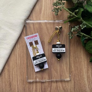 May include: A pair of earrings and a charm with the text "HELLO I'M READING. GO AWAY!". The earrings have a black and white design with gold-colored accents and chains. The charm has a gold-colored chain and a black and white design.