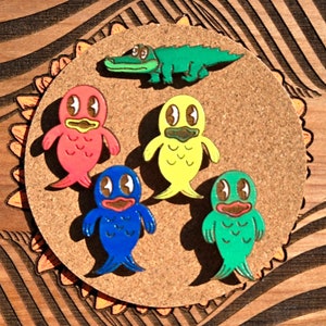 May include: Five enamel pins on a corkboard background. The pins are of a green alligator, a red fish, a yellow fish, a blue fish, and a green fish.