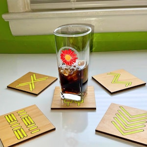 Peut inclure: Un grand verre orné d'une fleur rouge et d'un motif de crâne, rempli d'une boisson foncée, repose sur un dessous de verre en bois. Quatre dessous de verre en bois supplémentaires avec des motifs géométriques jaune néon entourent le verre.