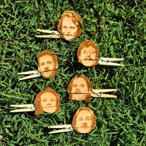 May include: Six wooden clothespins with different faces of people printed on them. The clothespins are arranged in a semi-circle on a bed of green grass.