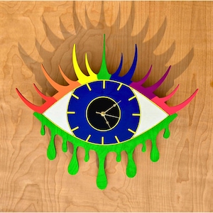 May include: A vibrant eye-shaped wall clock with a blue iris, white and gold details, and a black pupil. The clock features rainbow-coloured eyelashes and green dripping paint. It is mounted on a light brown wooden surface.