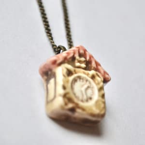 May include: A brown chain necklace with a ceramic cuckoo clock pendant. The clock is painted in shades of brown, white, and pink, and features a clock face with black hands.