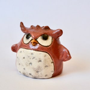May include: A small, ceramic owl figurine with a brown and white glaze. The owl has large, black eyes and a small, orange beak.