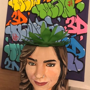 May include: A hand-painted planter shaped like a woman's face, with a green succulent plant on top. The planter is painted with realistic skin tones, brown hair, and a nose ring. A colorful graffiti art canvas is in the background.