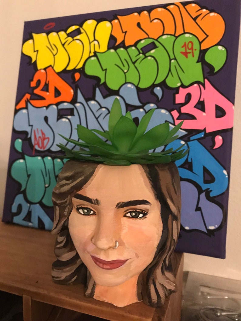 May include: A hand-painted planter shaped like a woman's face, with a green succulent plant on top. The planter is painted with realistic skin tones, brown hair, and a nose ring. A colorful graffiti art canvas is in the background.