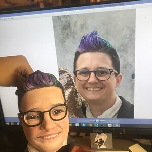 May include: A painted wooden head with glasses and purple and blue hair is held in front of a computer screen displaying a photo of a person with similar features. The head is a portrait, and the screen shows a smiling person.