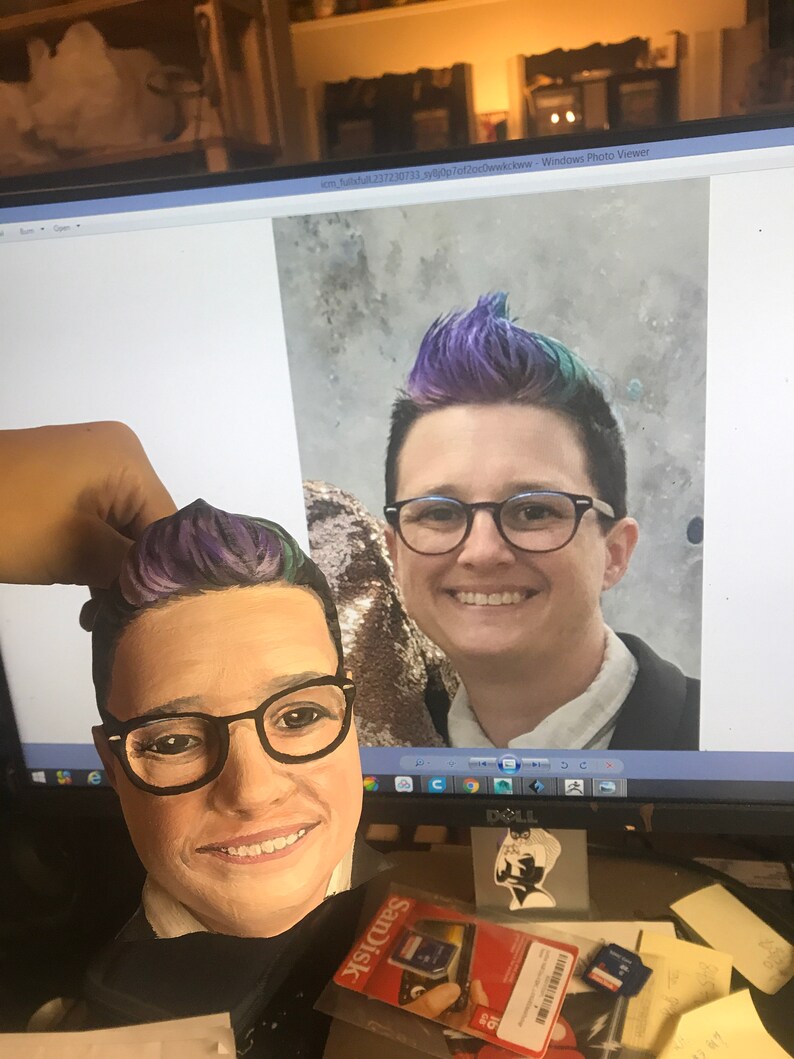 May include: A painted wooden head with glasses and purple and blue hair is held in front of a computer screen displaying a photo of a person with similar features. The head is a portrait, and the screen shows a smiling person.