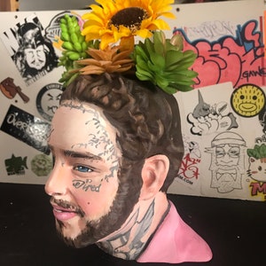 May include: A ceramic bust of a man with tattoos, styled as a planter. The bust is painted with realistic features and adorned with artificial sunflowers and succulents. The word "tired" is tattooed on the face. The bust is painted with a pink shirt.