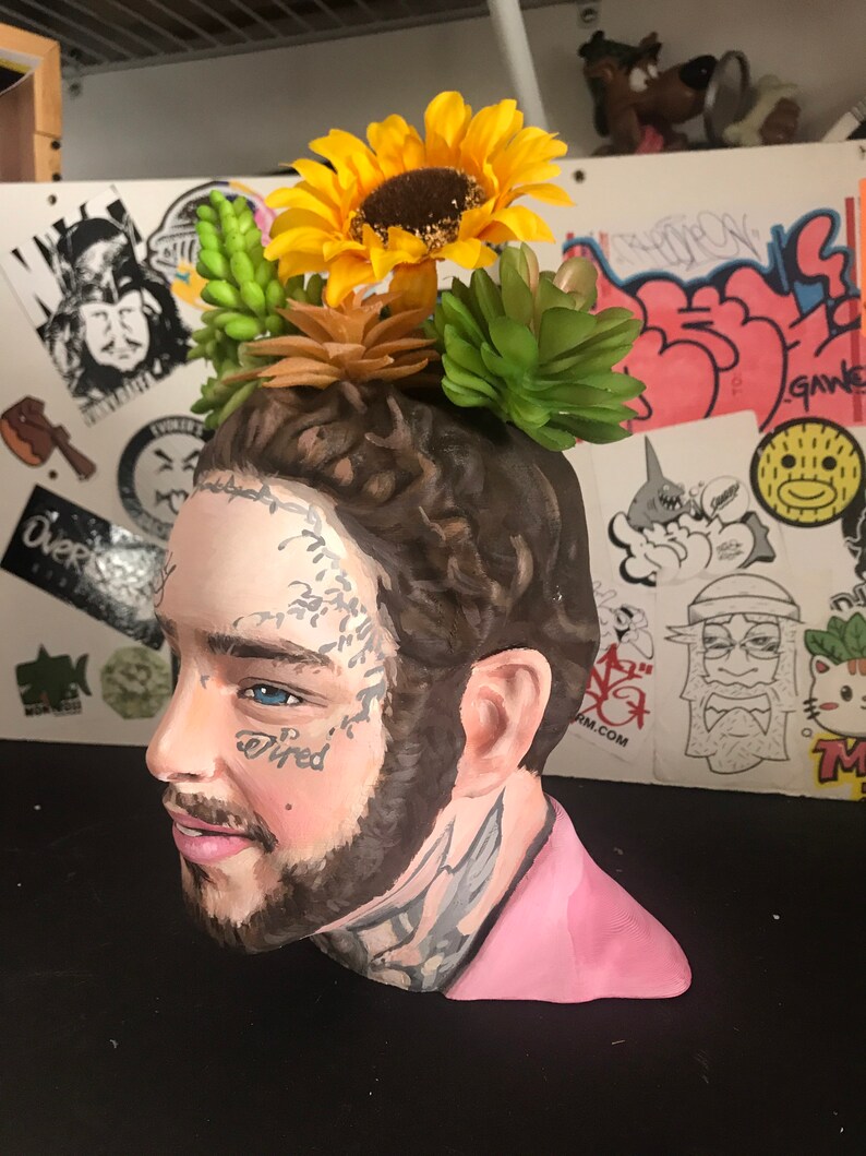 May include: A ceramic bust of a man with tattoos, styled as a planter. The bust is painted with realistic features and adorned with artificial sunflowers and succulents. The word "tired" is tattooed on the face. The bust is painted with a pink shirt.