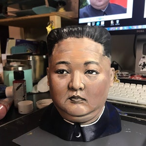 May include: A hand-painted bust of a man with dark hair and a stern expression, wearing a dark blue jacket with a white collar. The bust is painted with realistic skin tones and facial features. A computer monitor in the background displays a similar image.