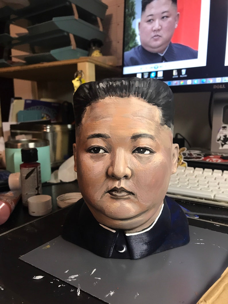 May include: A hand-painted bust of a man with dark hair and a stern expression, wearing a dark blue jacket with a white collar. The bust is painted with realistic skin tones and facial features. A computer monitor in the background displays a similar image.