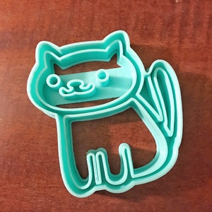 Pode incluir: Um cortador de biscoitos de pl&aacute;stico azul turquesa em forma de gato. O gato tem um design simples com um rosto listrado e uma cauda.