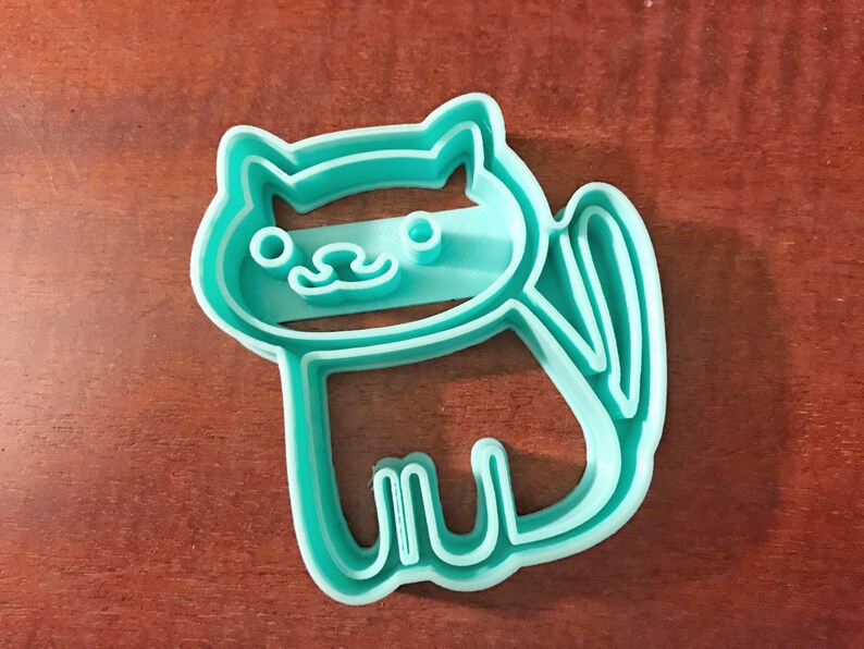 Pode incluir: Um cortador de biscoitos de pl&aacute;stico azul turquesa em forma de gato. O gato tem um design simples com um rosto listrado e uma cauda.