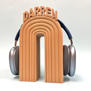 K&ouml;nnte beinhalten: Ein Holz-Kopfh&ouml;rerst&auml;nder mit dem Namen "DARREN" obenauf. Der St&auml;nder ist in Form eines Regenbogenbogens geformt und hat eine hellbraune Farbe. Der St&auml;nder h&auml;lt ein Paar silberne Kopfh&ouml;rer mit blauen Akzenten.