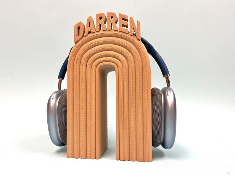 K&ouml;nnte beinhalten: Ein Holz-Kopfh&ouml;rerst&auml;nder mit dem Namen "DARREN" obenauf. Der St&auml;nder ist in Form eines Regenbogenbogens geformt und hat eine hellbraune Farbe. Der St&auml;nder h&auml;lt ein Paar silberne Kopfh&ouml;rer mit blauen Akzenten.