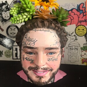 May include: A ceramic head planter featuring a painted portrait with tattoos and a beard. The planter is adorned with artificial sunflowers and succulents. The face has the words "Stay Away" and "Tired" tattooed on it.