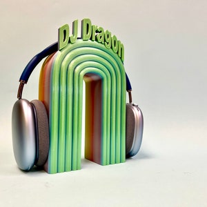 K&ouml;nnte beinhalten: Ein regenbogenfarbener Torbogen mit dem Text "DJ Dragon" oben, auf dem ein silberner Kopfh&ouml;rer liegt.