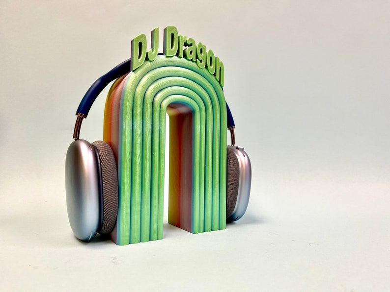 K&ouml;nnte beinhalten: Ein regenbogenfarbener Torbogen mit dem Text "DJ Dragon" oben, auf dem ein silberner Kopfh&ouml;rer liegt.