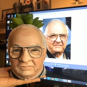 May include: A hand-painted ceramic planter shaped like a man's head, complete with glasses and a succulent plant on top. The planter is painted in realistic skin tones and features a black and white striped collar.