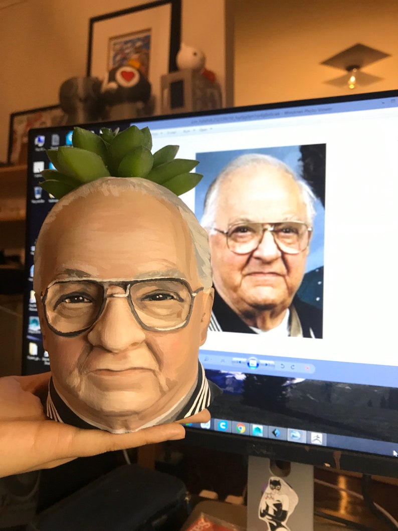 May include: A hand-painted ceramic planter shaped like a man's head, complete with glasses and a succulent plant on top. The planter is painted in realistic skin tones and features a black and white striped collar.
