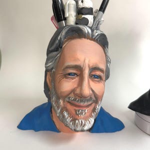 Puede incluir: Un portalápices pintado a mano con forma de cabeza de hombre, con ojos azules, pelo gris y barba. El portalápices contiene bolígrafos, rotuladores y una pequeña figura de panda. El hombre lleva una camisa azul.