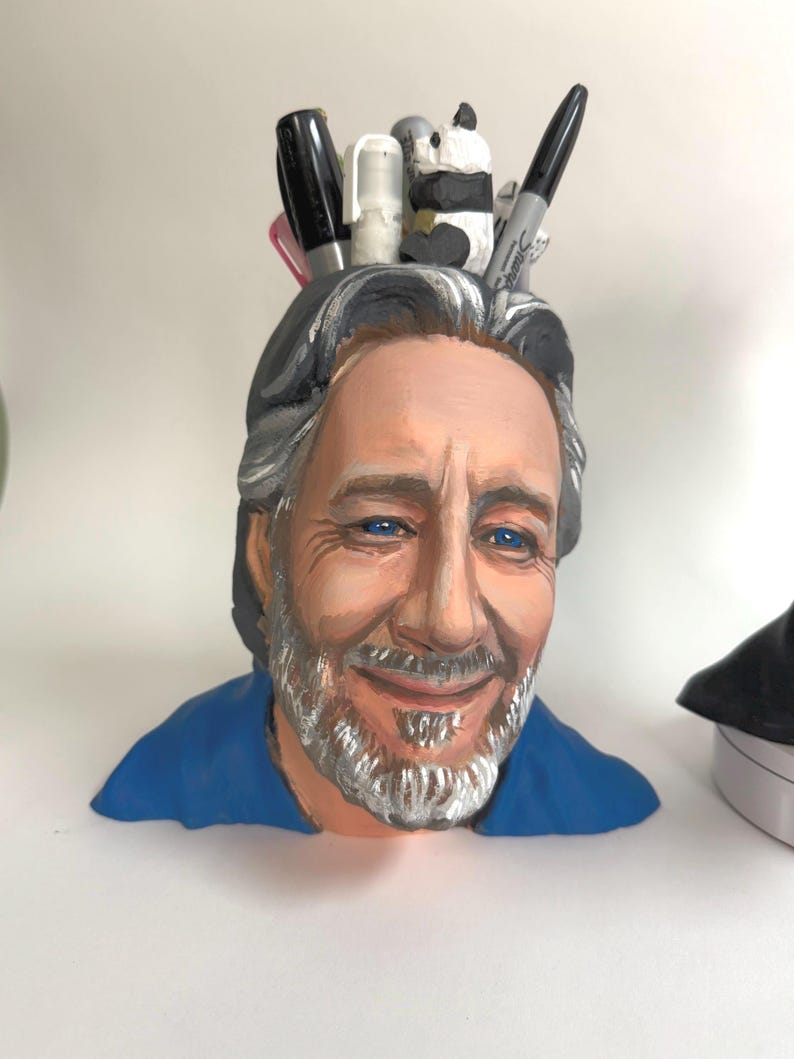 May include: A hand-painted pen holder shaped like a man's head, with blue eyes, gray hair, and a beard. The holder is filled with pens, markers, and a small panda figurine. The man is wearing a blue shirt.