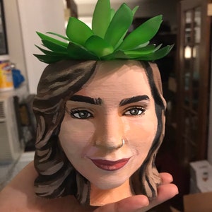 May include: A hand-painted planter shaped like a woman's face, with a green succulent plant on top. The face is painted with realistic features, including brown hair, dark eyes, and red lipstick. The planter is made of a light-colored material.