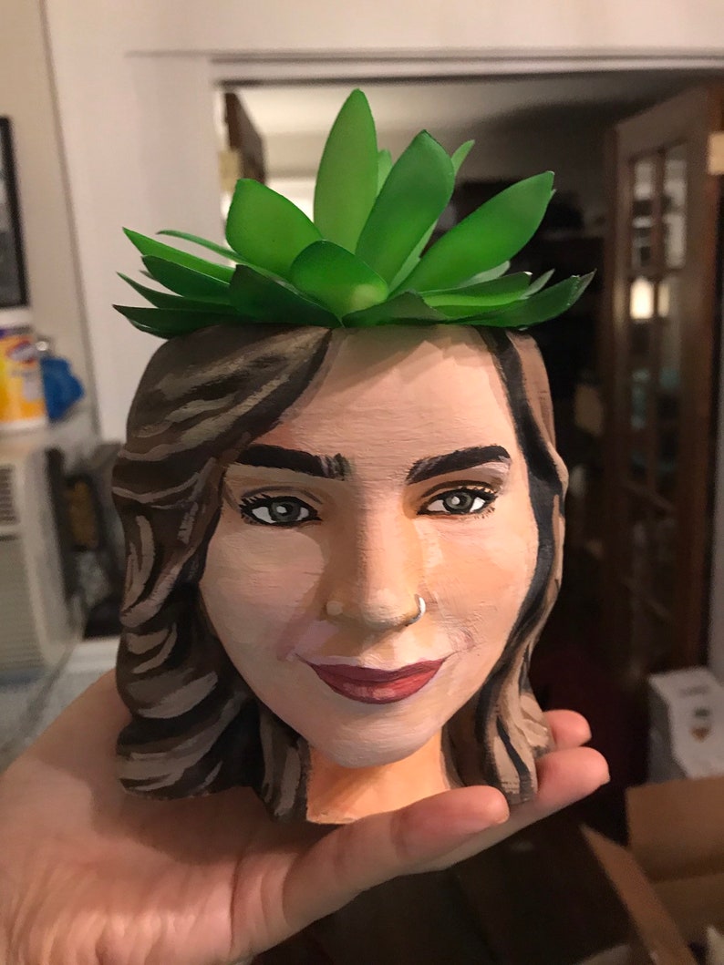 May include: A hand-painted planter shaped like a woman's face, with a green succulent plant on top. The face is painted with realistic features, including brown hair, dark eyes, and red lipstick. The planter is made of a light-colored material.