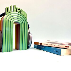 K&ouml;nnte beinhalten: Ein regenbogenfarbener 3D-gedruckter Kopfh&ouml;rerst&auml;nder mit dem Text "DJ Dragon" oben. Der St&auml;nder h&auml;lt ein Paar silberne Kopfh&ouml;rer. Zwei B&uuml;cher sind auf einem Tisch vor dem St&auml;nder gestapelt. Das obere Buch tr&auml;gt den Titel "Seven Taoist Masters" und das untere Buch den Titel "The Journey to the West."