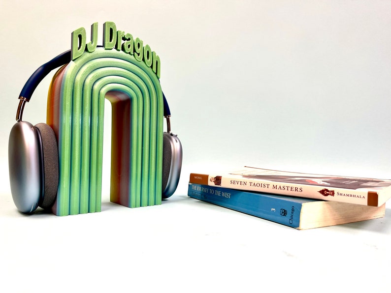K&ouml;nnte beinhalten: Ein regenbogenfarbener 3D-gedruckter Kopfh&ouml;rerst&auml;nder mit dem Text "DJ Dragon" oben. Der St&auml;nder h&auml;lt ein Paar silberne Kopfh&ouml;rer. Zwei B&uuml;cher sind auf einem Tisch vor dem St&auml;nder gestapelt. Das obere Buch tr&auml;gt den Titel "Seven Taoist Masters" und das untere Buch den Titel "The Journey to the West."