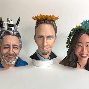 May include: Three ceramic busts with realistic facial features. The first bust has grey hair and a beard, with pens and a panda figurine on top. The second bust has a yellow flower crown. The third bust has a succulent crown.