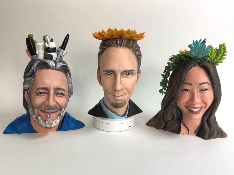 May include: Three ceramic busts with realistic facial features. The first bust has gray hair and a beard, with pens and a panda figurine on top. The second bust has a yellow flower crown. The third bust has a succulent crown.