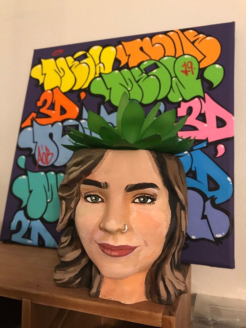 May include: A hand-painted ceramic planter shaped like a woman's face, with a green paper succulent on top. The planter is brown and tan with detailed facial features. A purple canvas with colorful graffiti art is in the background.