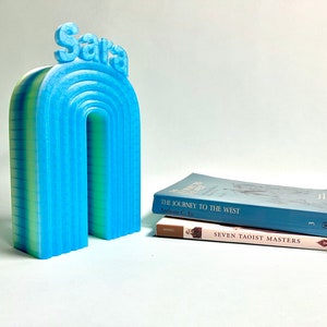 K&ouml;nnte beinhalten: Ein blauer und gr&uuml;ner 3D-gedruckter Regenbogen-Buchst&uuml;tze mit dem Namen "Sara" oben. Der Buchst&uuml;tze h&auml;lt zwei B&uuml;cher, "Die Reise nach Westen" und "Sieben Taoistische Meister".