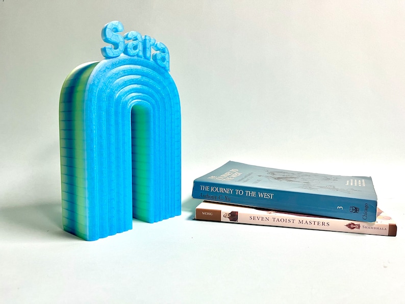 K&ouml;nnte beinhalten: Ein blauer und gr&uuml;ner 3D-gedruckter Regenbogen-Buchst&uuml;tze mit dem Namen "Sara" oben. Der Buchst&uuml;tze h&auml;lt zwei B&uuml;cher, "Die Reise nach Westen" und "Sieben Taoistische Meister".