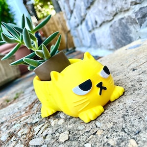 Peut inclure: Un pot de fleurs en forme de chat jaune avec une expression boudeuse. Le pot est en plastique et contient une petite plante succulente.