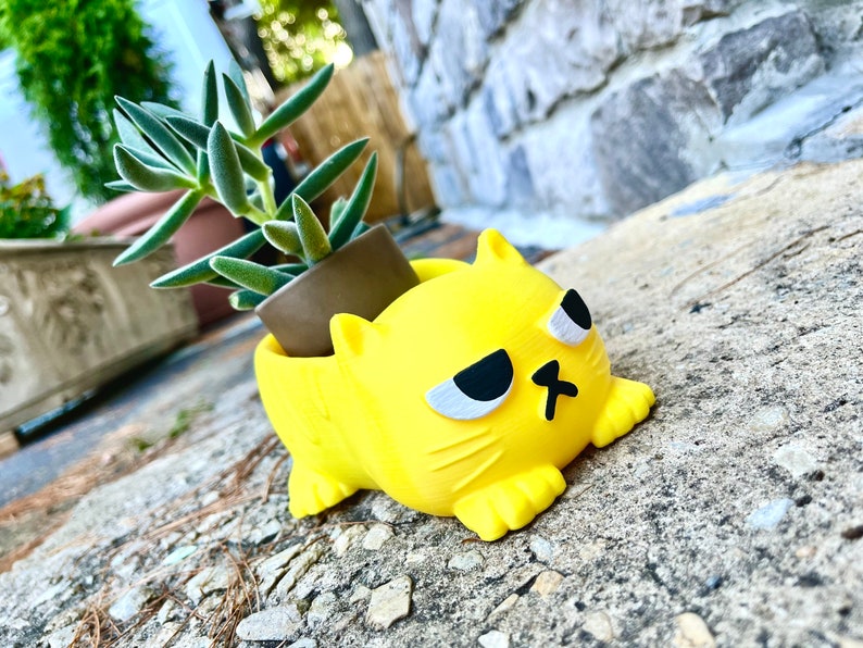 以下が含まれることがあります： 不機嫌そうな表情をした黄色い猫の形をしたプランター。プランターはプラスチック製で、中に小さな多肉植物が植えられています。