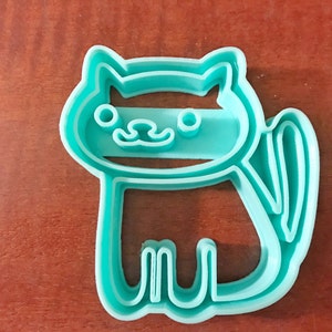 Pode incluir: Um cortador de biscoitos de pl&aacute;stico azul turquesa em forma de gato com um rosto simples e uma cauda.