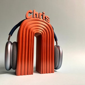 K&ouml;nnte beinhalten: Ein Kopfh&ouml;rerst&auml;nder aus Holz mit dem Wort "Chris" oben drauf. Der St&auml;nder ist orange und hat eine geschwungene Form. Ein silberner Kopfh&ouml;rer liegt auf dem St&auml;nder.