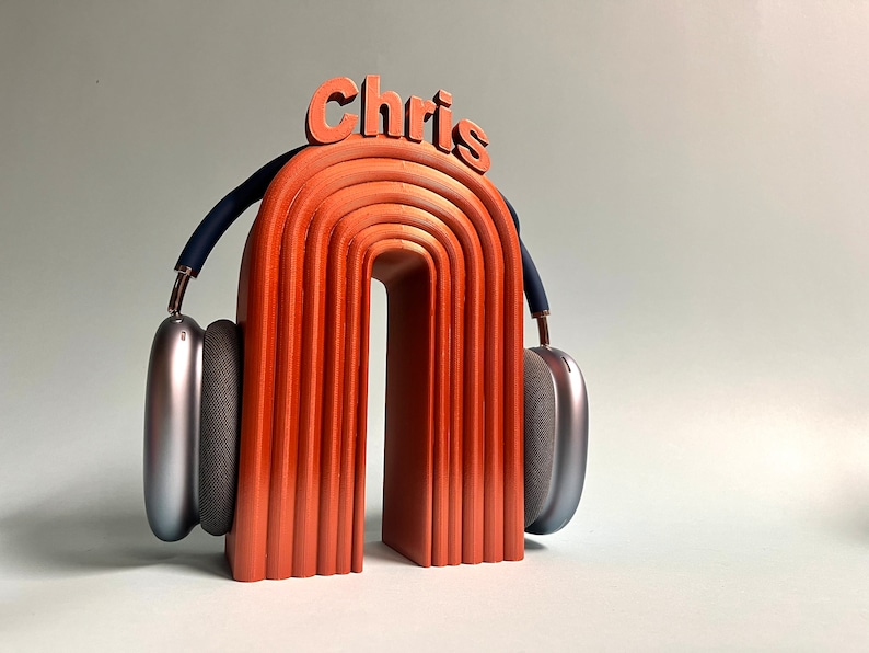 K&ouml;nnte beinhalten: Ein Kopfh&ouml;rerst&auml;nder aus Holz mit dem Wort "Chris" oben drauf. Der St&auml;nder ist orange und hat eine geschwungene Form. Ein silberner Kopfh&ouml;rer liegt auf dem St&auml;nder.