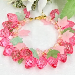 Peut inclure: Un bracelet charmant avec des breloques fraises roses translucides, des perles roses et transparentes, et des accents de feuilles. Le bracelet est sécurisé par un fermoir doré. Les breloques fraises mesurent environ 2 cm de long.