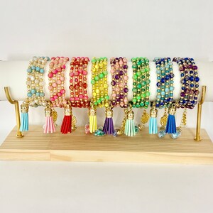 Peut inclure: Un présentoir de bracelets en perles colorées avec des accents dorés et des pompons. Les bracelets sont disposés sur un support blanc avec des accents dorés.