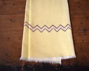 May include: A yellow kitchen towel with a white and brown zig-zag pattern along the bottom edge. The towel has a fringe border.