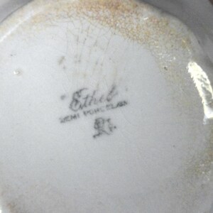 May include: Close-up of a white semi-porcelain dish with a scalloped edge. The bottom of the dish features the name "Ethel" in script, along with "SEMI PORCELAIN" and a small emblem. The rim has a faded gold-colored trim.