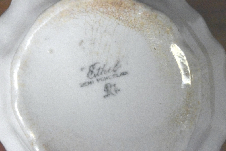 May include: Close-up of a white semi-porcelain dish with a scalloped edge. The bottom of the dish features the name "Ethel" in script, along with "SEMI PORCELAIN" and a small emblem. The rim has a faded gold-colored trim.