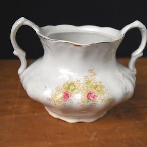 May include: White ceramic sugar bowl with two handles and a floral design featuring pink and yellow roses. The rim has a gold trim. The bowl sits on a wooden surface.