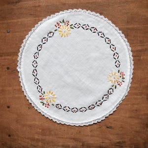 May include: A white, round doily with a delicate lace edge. The center features embroidered floral designs in yellow, red, and green, framed by a black and pink geometric border. The doily is placed on a wooden surface.