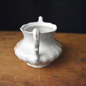 May include: White ceramic creamer with a handle and a small spout. The creamer has a scalloped edge and is decorated with floral patterns in pink, yellow, and green. The handle has a gold trim.