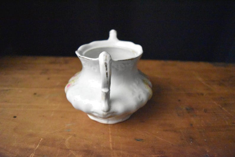 May include: White ceramic creamer with a handle and a small spout. The creamer has a scalloped edge and is decorated with floral patterns in pink, yellow, and green. The handle has a gold trim.