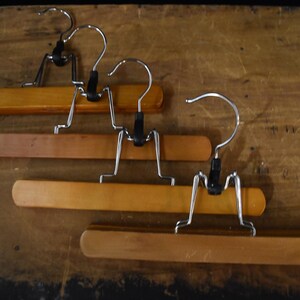 May include: Four wooden pant hangers with smooth, light to medium brown finishes. Each hanger features a silver-toned metal hook, a black plastic swivel connector, and a metal clamping mechanism designed to hold garments securely in place.